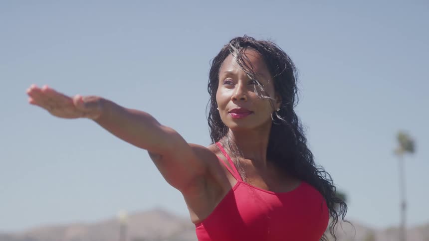 A woman stands under the sun wearing a red dress and red lipstick stretching her right arm forward