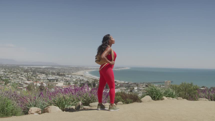 A woman in a red athletic top and leggings stands on a hill overlooking a city and the ocean she is looking out at the scenery with her hands on her hips taking in the view
