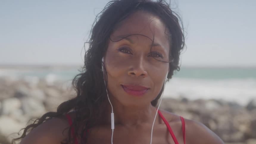 Woman in red bathing suit smiles warmly at the beach with earphones in