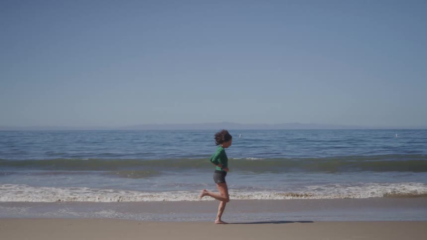 A woman wearing a long sleeved green shirt and black shorts runs barefoot on a sandy beach next to the ocean