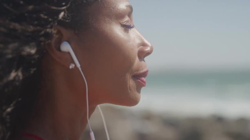 Woman with closed eyes wearing earphones with the ocean in the background on a sunny day