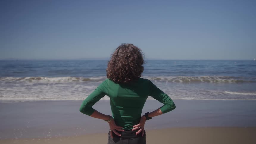 The video shows a woman standing with her back to the camera looking at the ocean she is wearing a green longsleeved shirt and black shorts with her hands on her hips
