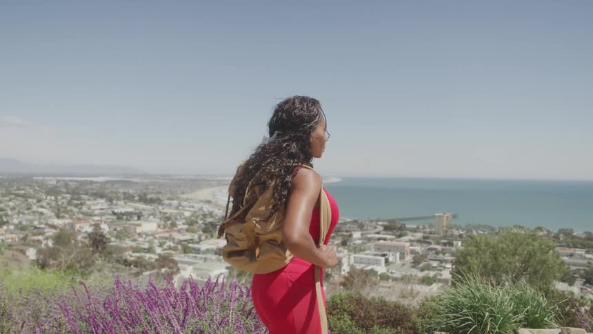 A woman wearing a red dress and a tan backpack stands on a hill overlooking a coastal city and the ocean on a sunny day