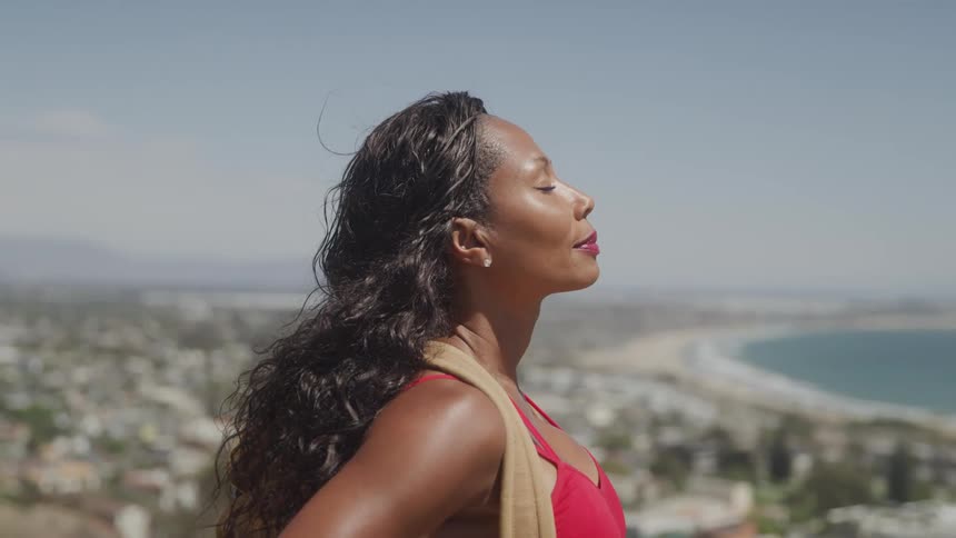 A woman with her eyes closed and long dark wavy hair stands on a hill overlooking the ocean and a coastal town