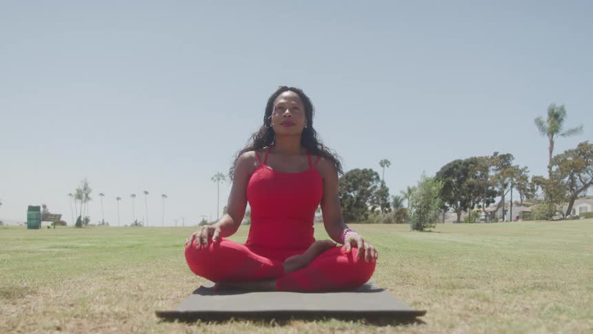 A woman sits crosslegged on a yoga mat in a grassy outdoor park