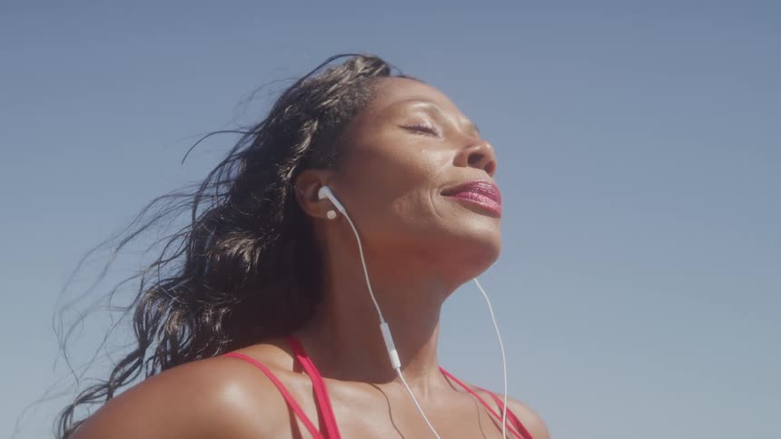 The video shows a closeup of a woman with her eyes closed wearing a red top and white wired earphones her head is tilted back and she appears to be enjoying the moment with the wind gently blowing her dark hair against a clear blue sky