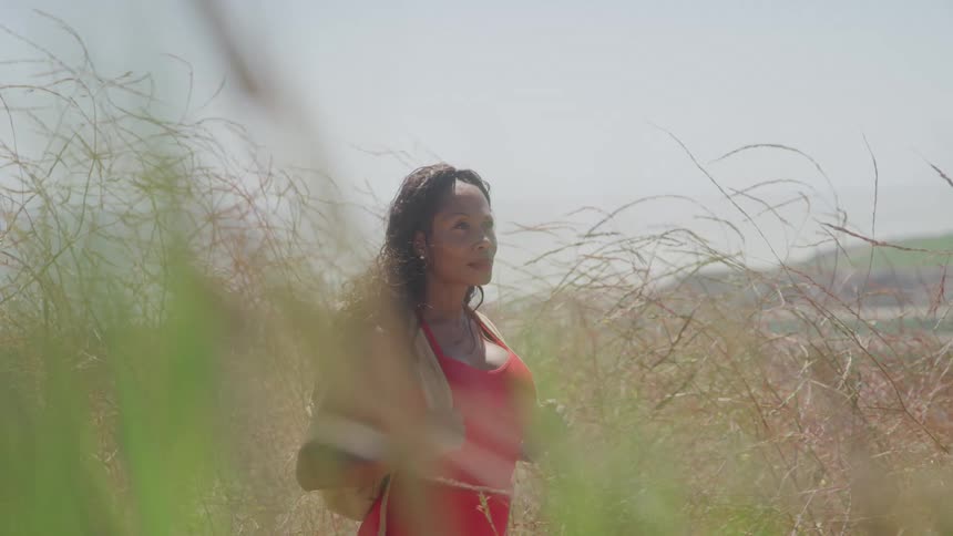 A woman in a red tank top with a backpack stands in a field of tall dry grass on a bright hazy day