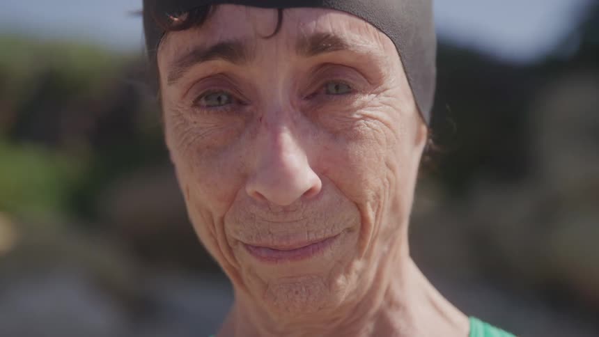 The video shows a closeup of a middleaged woman with a swimming cap and goggles on her head looking at the camera and smiling her face shows some wrinkles suggesting she is in her mid50s or older
