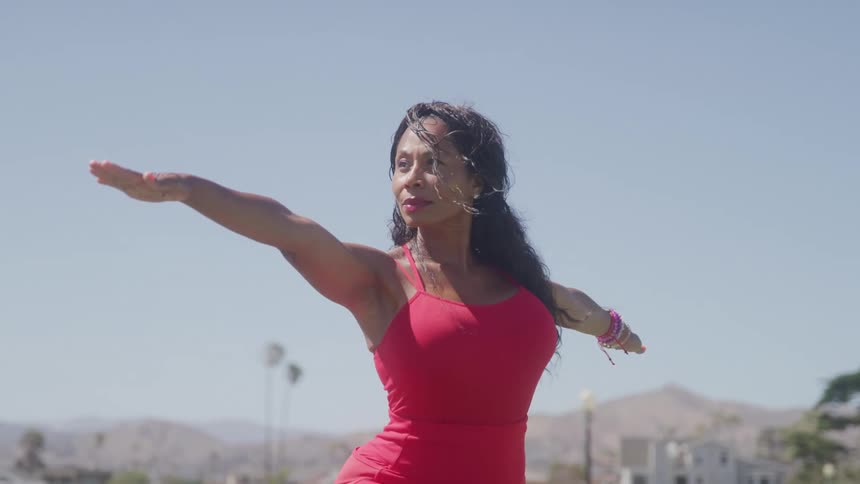 A woman with long dark hair and a snug red top is doing yoga outdoors with her arms stretched out to the side