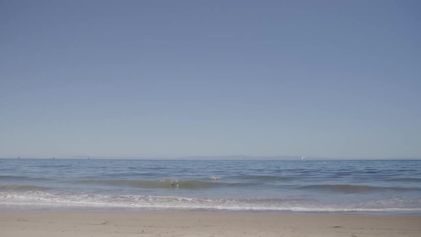 Gentle waves roll onto a quiet sandy beach under a clear blue sky with distant sailboats on the horizon