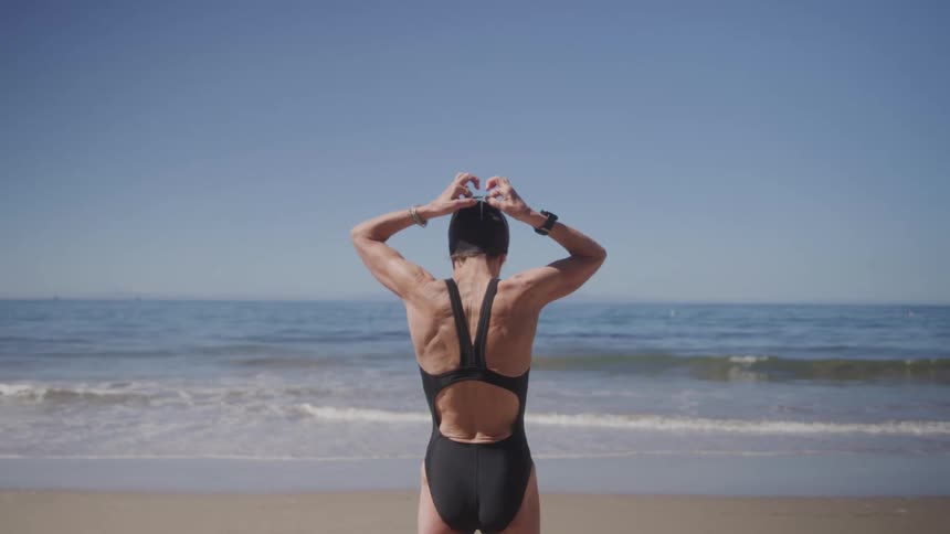 Woman in a black swimsuit adjusts her swimming goggles facing the sea