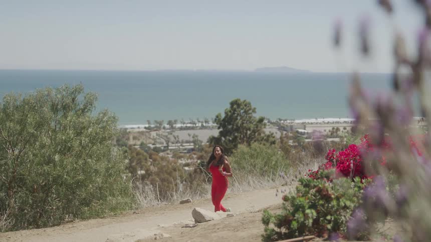 A woman in a bright red outfit walks along a scenic hillside trail with ocean views and vibrant flowers