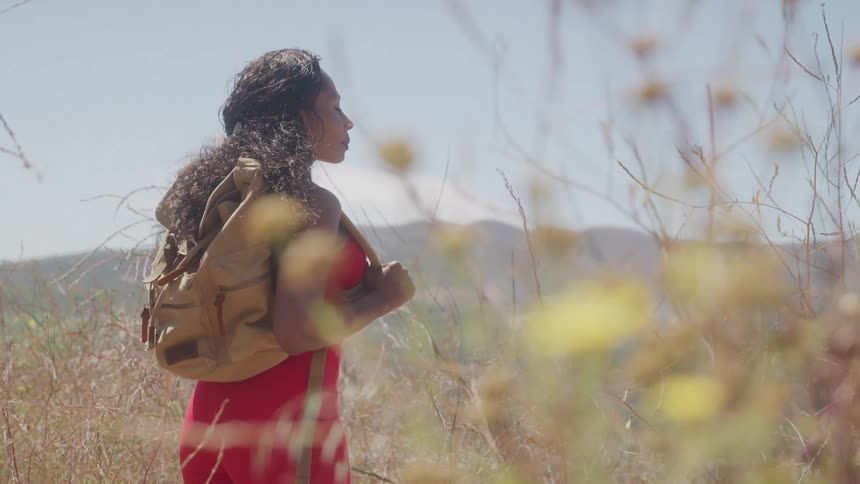 The video shows a woman with a backpack walking through a field of tall dry grass she is wearing a red dress and is looking off to the side enjoying the sunny day