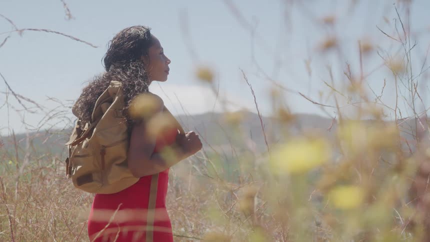 Woman with a backpack hikes along a dry trail surrounded by vegetation under strong sunlight