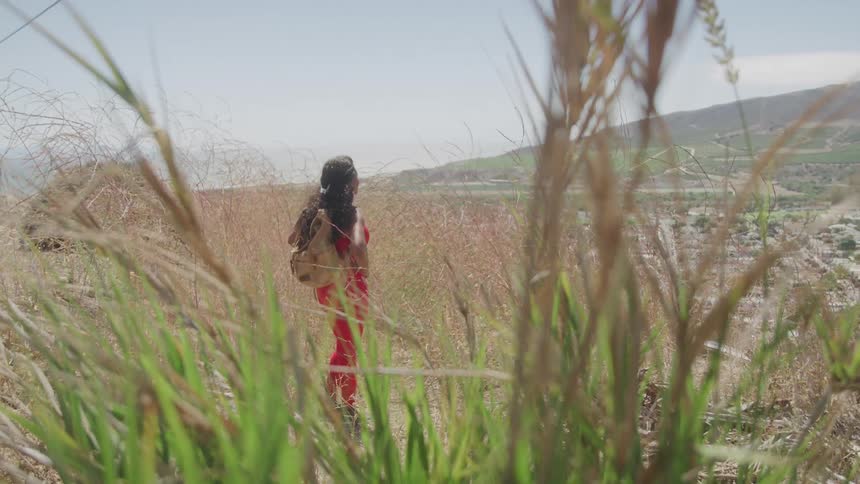 A woman wearing red trousers stands on a hill on the outskirts of town looking down at the city at midday surrounded by grass