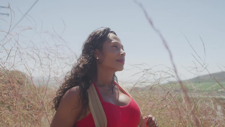 The video shows a woman with long curly brown hair wearing a red sports bra standing in a field of tall dry grass she is looking off to her right and a backpack strap is visible on her shoulder