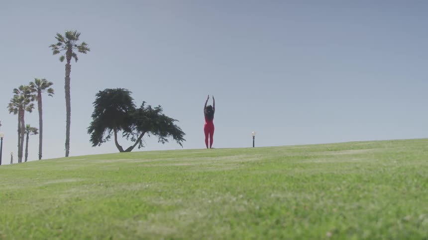 Woman in red outfit practices a yoga pose on a grassy field with palm trees in the background