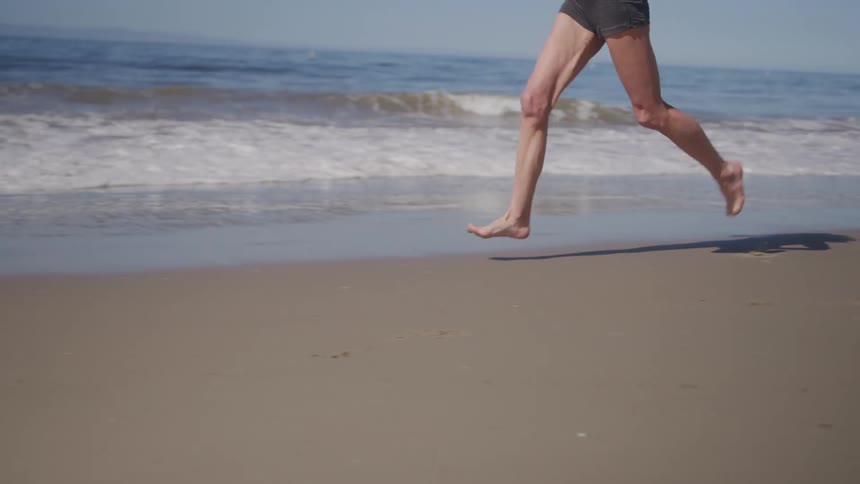 The video shows a lowangle shot of a person jogging barefoot on a sandy beach the ocean waves are visible in the background crashing gently on the shore under a clear sunny sky