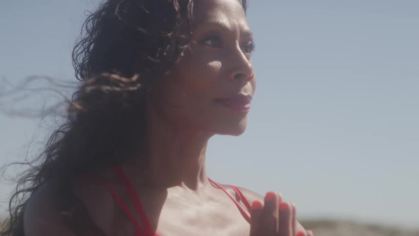 The video shows a woman with dark curly hair and a red bikini top standing outdoors in the sun she is looking up with her eyes closed as if meditating or soaking in the sunlight
