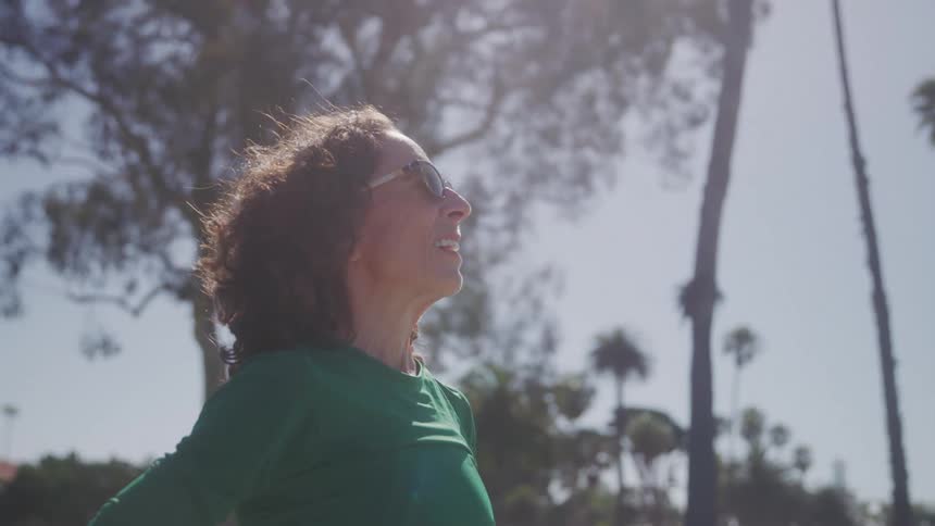 A middleaged woman with curly hair and sunglasses wearing a green shirt stands outdoors on a sunny day looking up with a content expression as sunlight filters through the trees