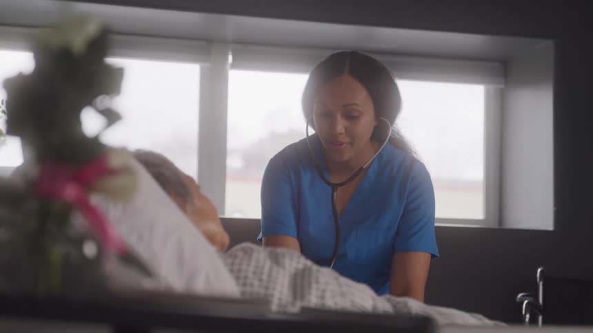 A nurse wearing blue scrubs and a stethoscope leans over a patient who is lying in a hospital bed