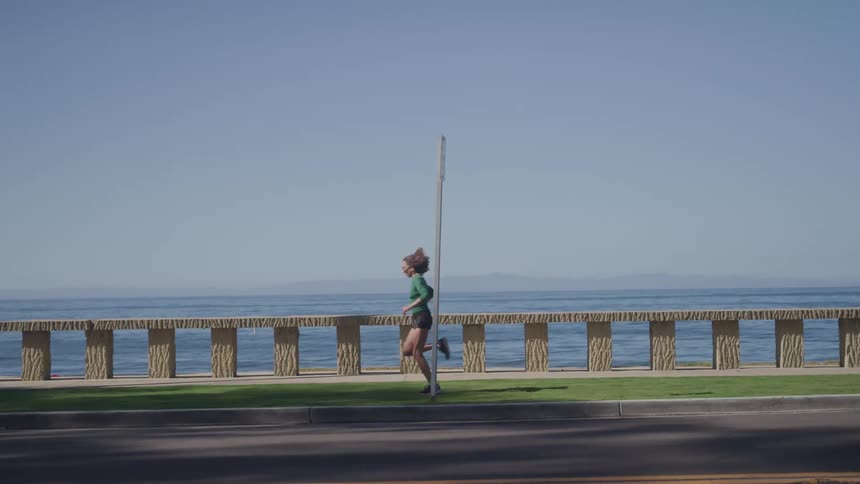 The video shows a jogger running on a paved path next to the ocean on a bright sunny day the runner is seen in profile wearing a green longsleeved shirt and black shorts with a calm wide ocean behind them