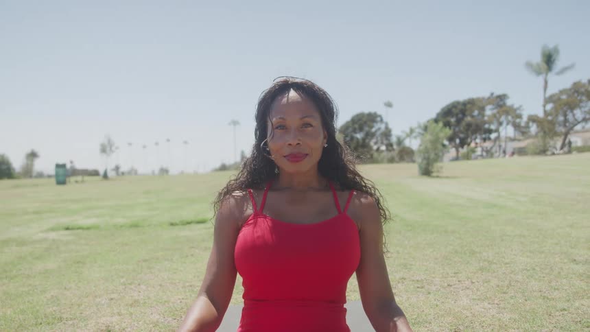 The video shows a woman with long dark hair wearing a red sports bra standing in a sunny grassy field she is looking down at the ground then looks up and smiles at the camera