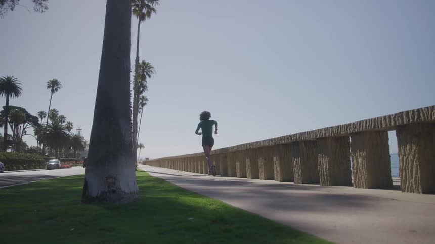 The video shows a woman with curly hair running along a paved walkway next to the ocean the sun is shining brightly and there are palm trees and a car on the street in the background