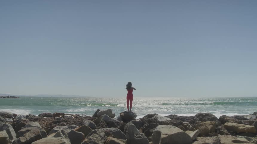 A woman in a red jumpsuit stands on a rocky shoreline facing the ocean with her hands in a prayer pose as waves crash behind her