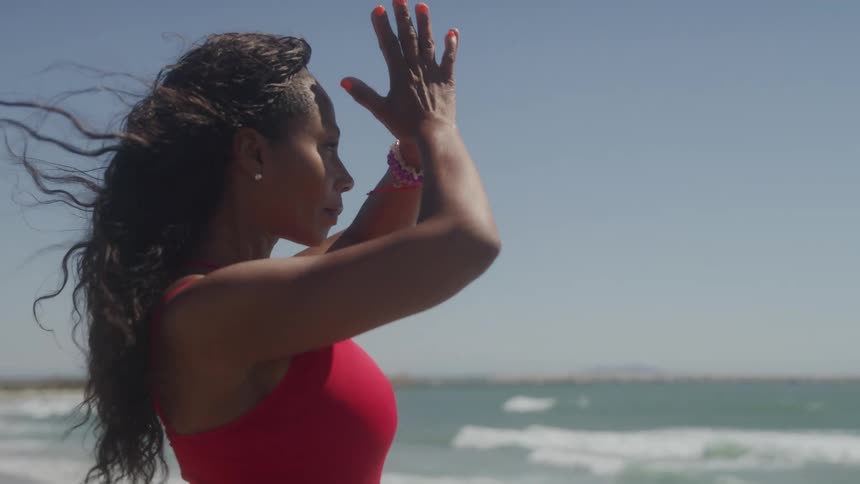 A woman in a form fitting red top stands on a shore on a sunny day and raises her hands above her head before bringing them together in a prayer position
