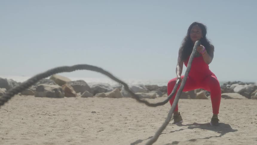 A woman in a red jumpsuit performs a squat while vigorously swinging heavy ropes as part of an intense workout on a sandy beach