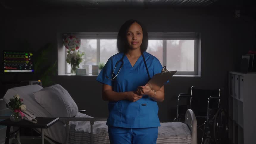 A steady eyelevel shot of a female healthcare professional in blue scrubs standing in a dimly lit hospital room holding a clipboard and looking directly at the camera with a serious expression