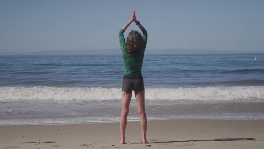 A woman with curly hair stands barefoot on a sandy beach stretching her arms out to her sides and then raising them over her head