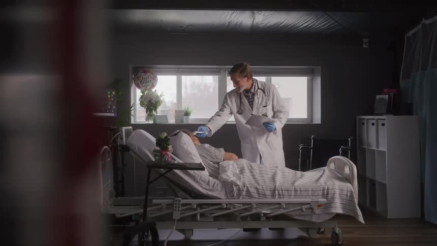 As the camera slowly pans right to reveal the scene a doctor in a white coat tends to a patient resting in a hospital bed