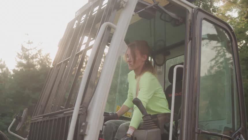 A woman with her long hair in a ponytail sits inside the cab of a large piece of construction equipment wearing a light green shirt and black gloves while operating the controls