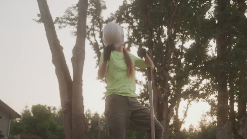 A woman in work clothes wipes her brow while holding a helmet after shoveling dirt