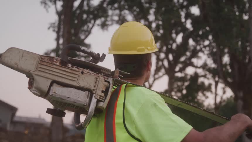 A construction worker carries a chainsaw in the woods wearing a hard hat and a green vest at midday