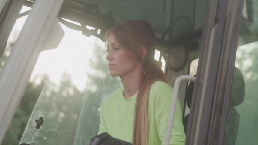 The video shows a young woman with long hair wearing a light green shirt and black gloves sitting inside the cab of a large piece of equipment she is seen looking to the side appearing focused and serious as she operates the machinery