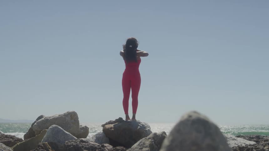 A woman wearing a snug red top and leggings stands on a rock on a sunny day with her back to the ocean raising her hands over her head and then bringing them down to her chest in a prayer position