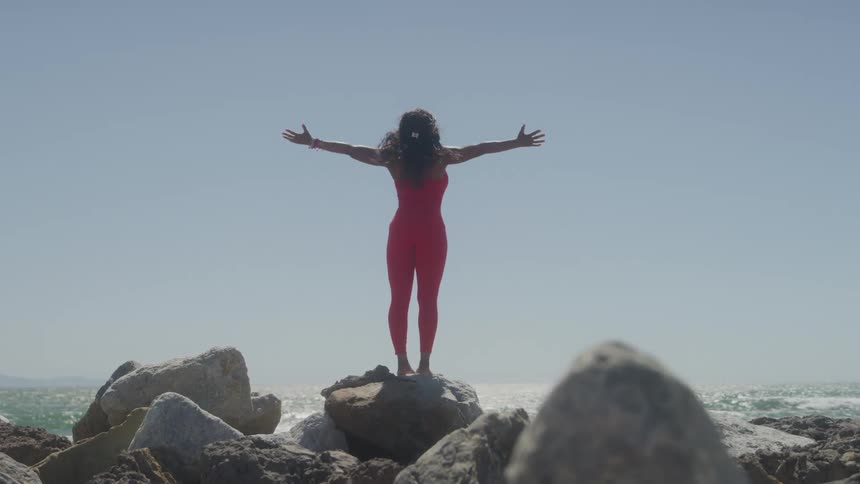 Woman in a red jumpsuit raises her arms facing the sea on the rocks