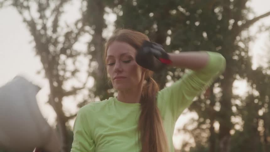 A young woman in a field surrounded by trees at midday removes her hard hat to wipe sweat from her face then puts it back on and picks up a shovel to begin digging the soil