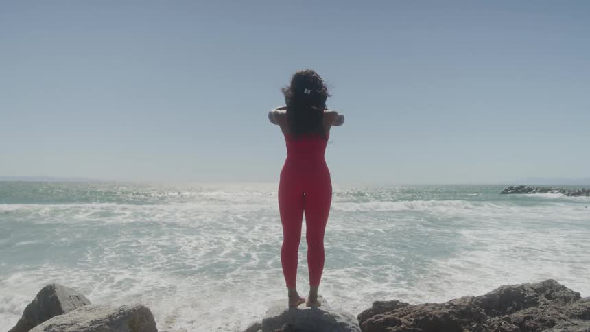 A woman in a snug red top and leggings stands on a rock on a sunny day with her back to the ocean raising her hands over her head and then bringing them down to her chest in a prayer position