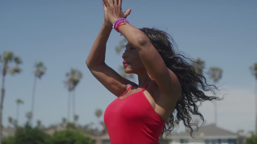 A woman wearing a red top and purple bracelets raises her arms and brings her hands together in a prayer position while doing yoga outdoors on a sunny day