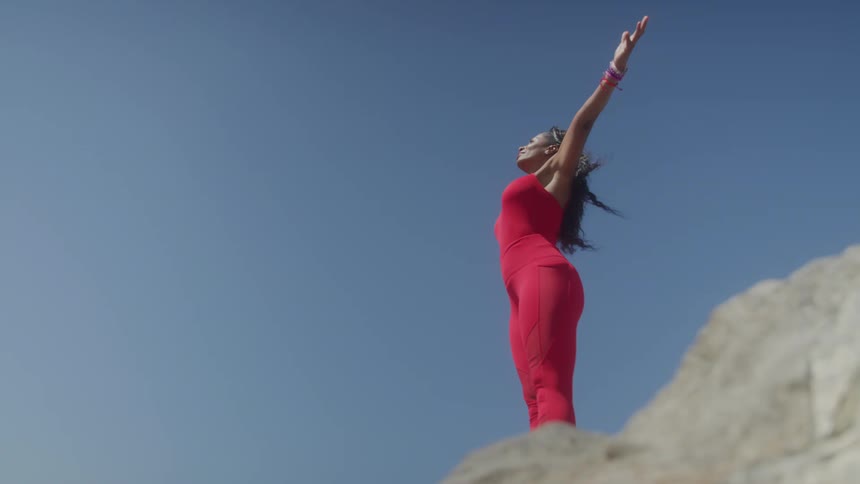 The video shows a woman standing on a rock with a clear blue sky in the background she is wearing a red athletic outfit and raises her arms up with her palms open as if celebrating a victory or enjoying the moment