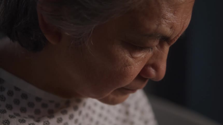 A closeup shot of an older person with graying hair and deep wrinkles on their face the person is looking down with a serious expression and they are wearing what appears to be a hospital gown