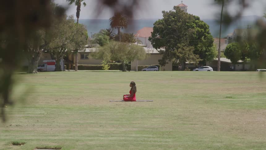 A woman wearing a bright red sleeveless dress sits on a red yoga mat in a lotus position in the middle of a large grassy field