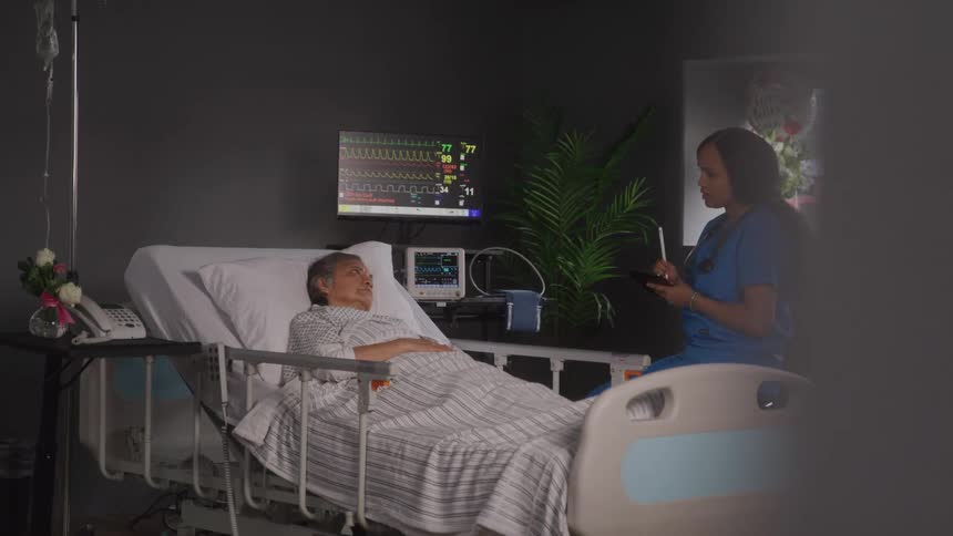 The video shows an elderly patient lying in a hospital bed with an iv drip and a heart rate monitor next to them the patient appears to be looking to their right and a person is visible in the foreground but is out of focus