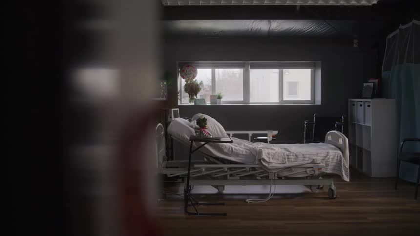 A hospital bed in a dark quiet room is lit gently by natural light from a window