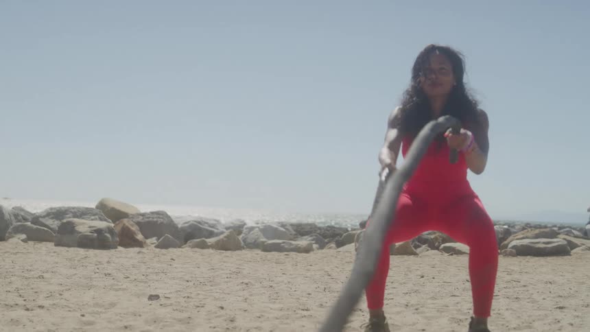 A woman in bright red workout clothes trains with battle ropes on a sandy beach near the ocean