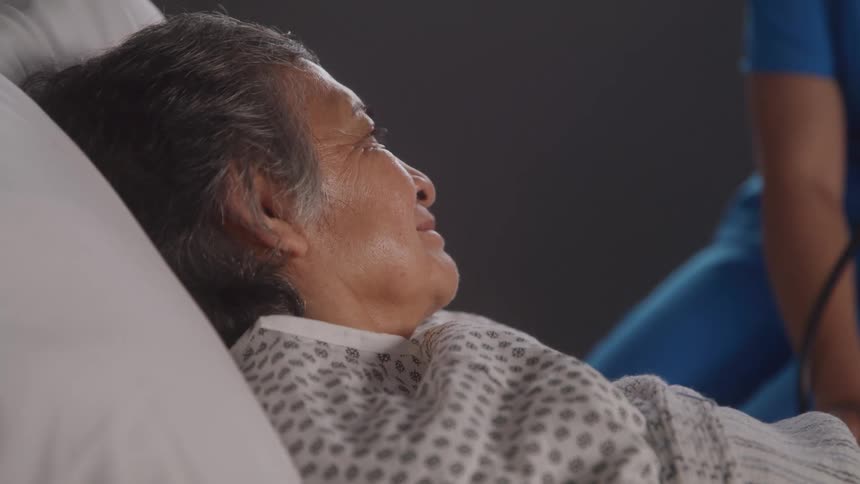 The video shows an elderly patient lying in a hospital bed looking away from the camera a medical professional in a blue shirt is visible in the background seeming to attend to the patient